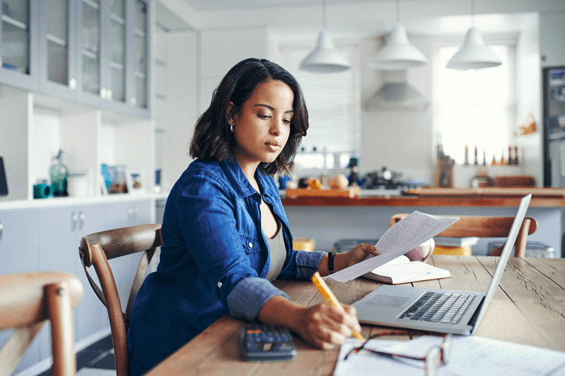 a woman sits at a kitchen table in front of a laptop writing on a piece of paper